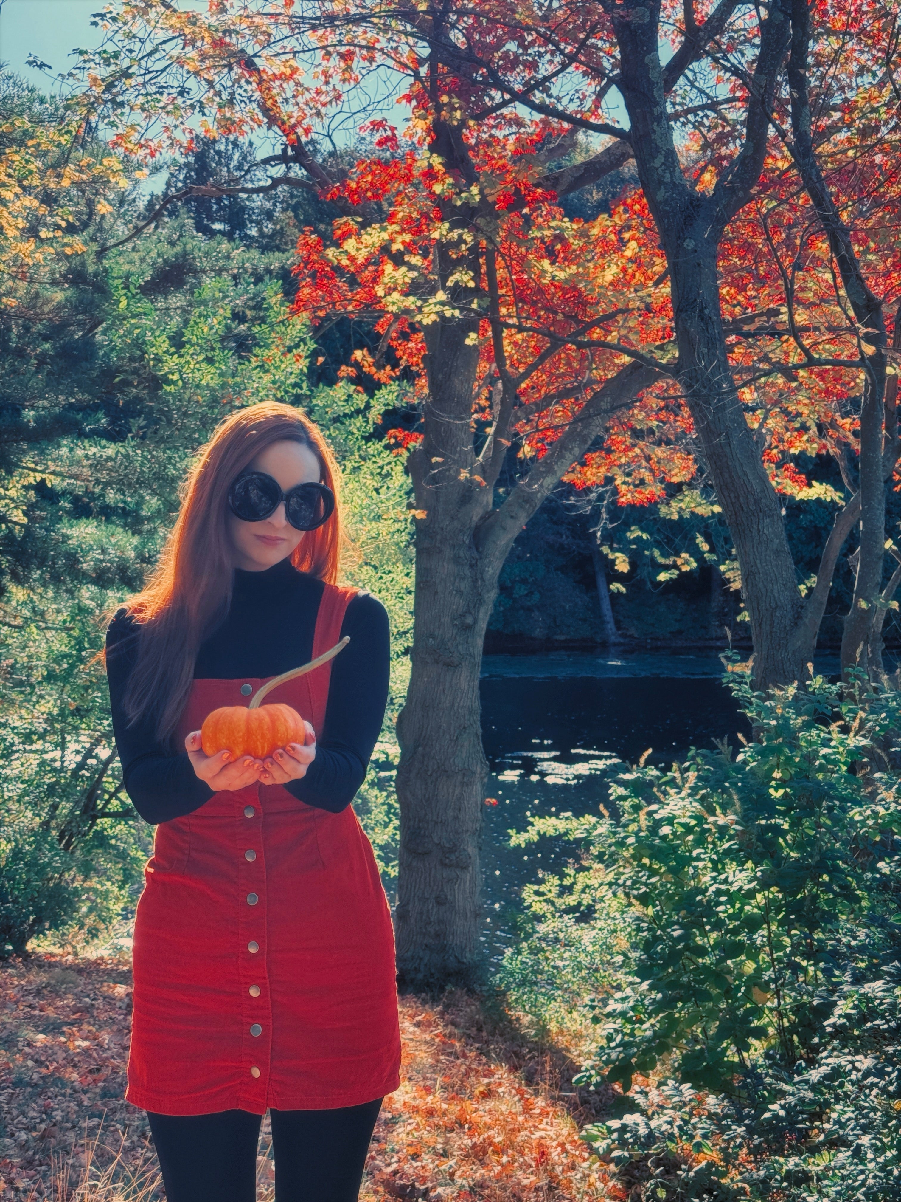 Redheaded woman holding out a small pumpking wearing an orange overalls dress with a black turtleneck.